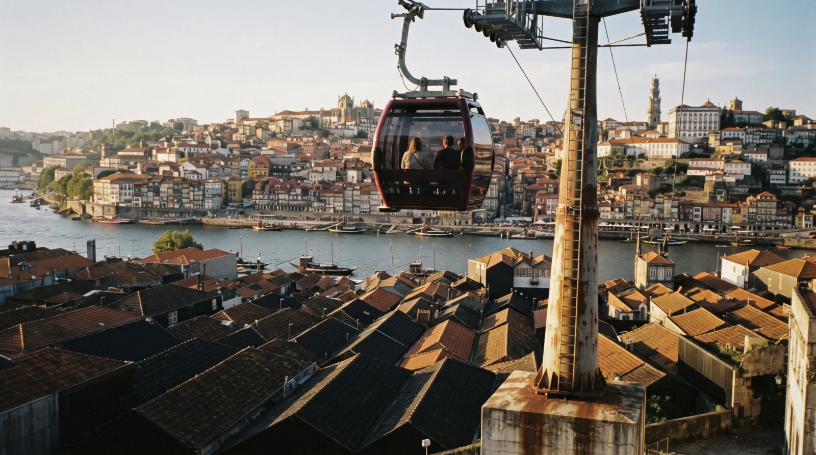 A red-framed Gaia cable car cabin crossing above the port lodge roofs of Vila Nova de Gaia at 18:20 with the Douro and the Porto bank behind it, two passengers small and seen from behind, gaia cable car