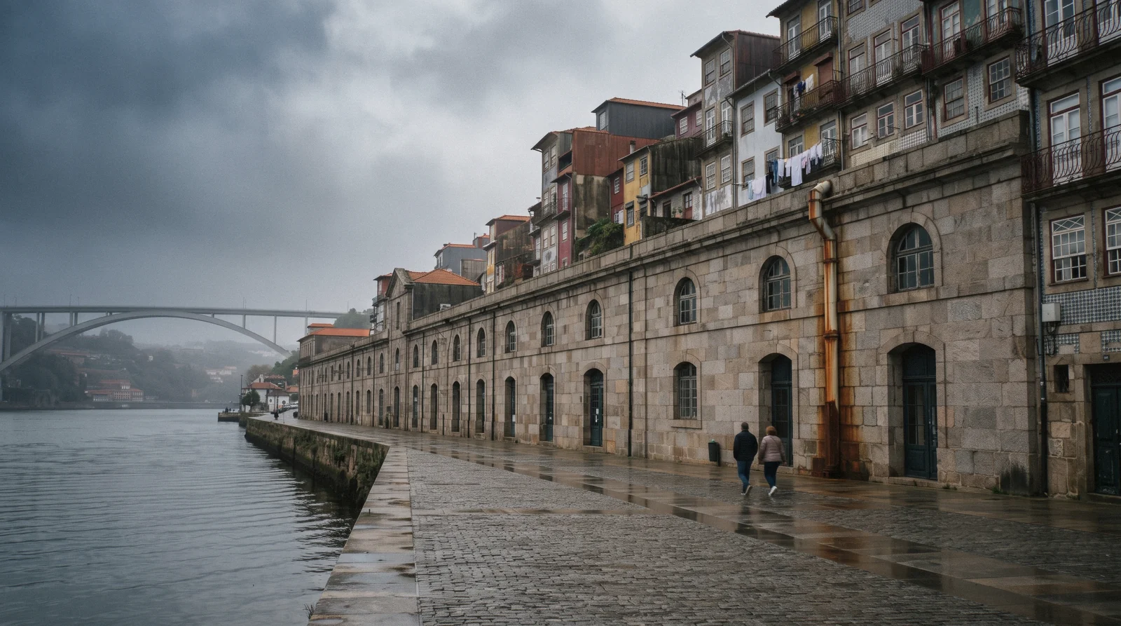 The long granite facade of the Alfandega customs house on the Douro at 09:10 with the Arrabida bridge beyond and washing on the balconies of the Miragaia lanes above, Miragaia Porto