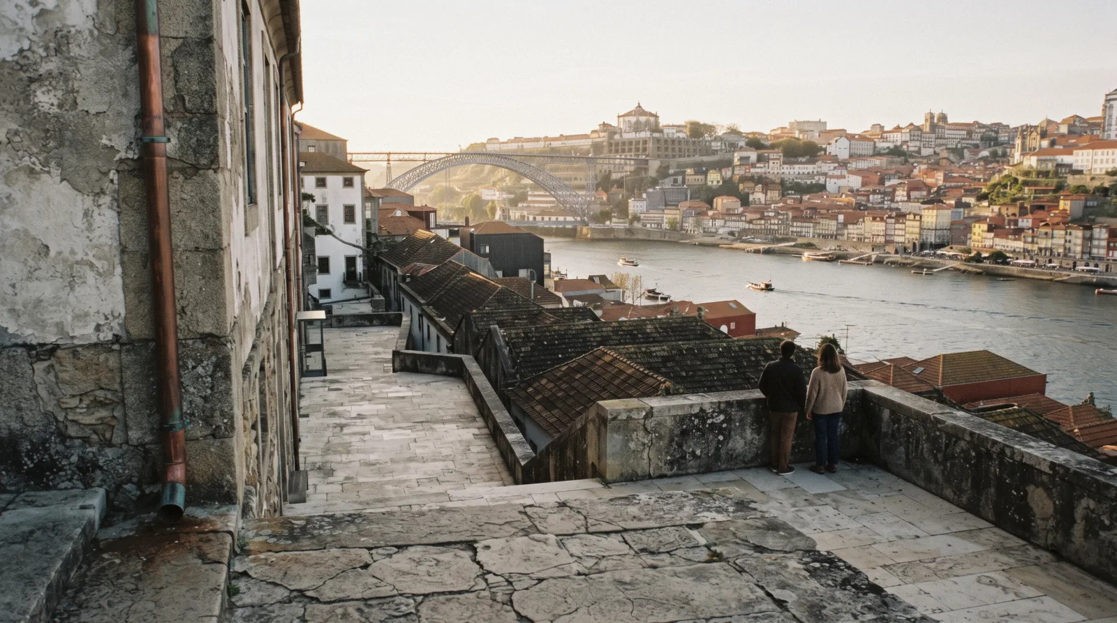 The pale stone terraces and courtyards of the WOW museum quarter above the port lodges in Gaia at 17:30, the Douro and the Porto bank below, two visitors small and seen from behind at a parapet, world of wine porto